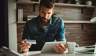 A man sitting at a table, holding a tablet and his debit card looking thoughtful