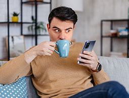 Man sitting on a sofa drinking from a mug looking at his phone